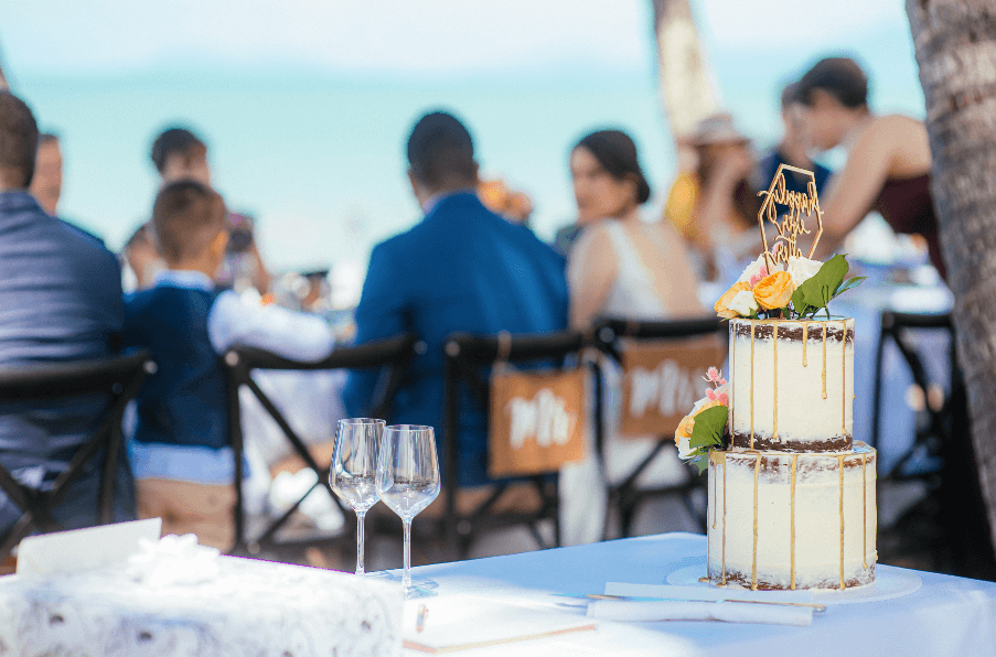 wedding cake and wine glasses on foreground, bride and groom with guests on background