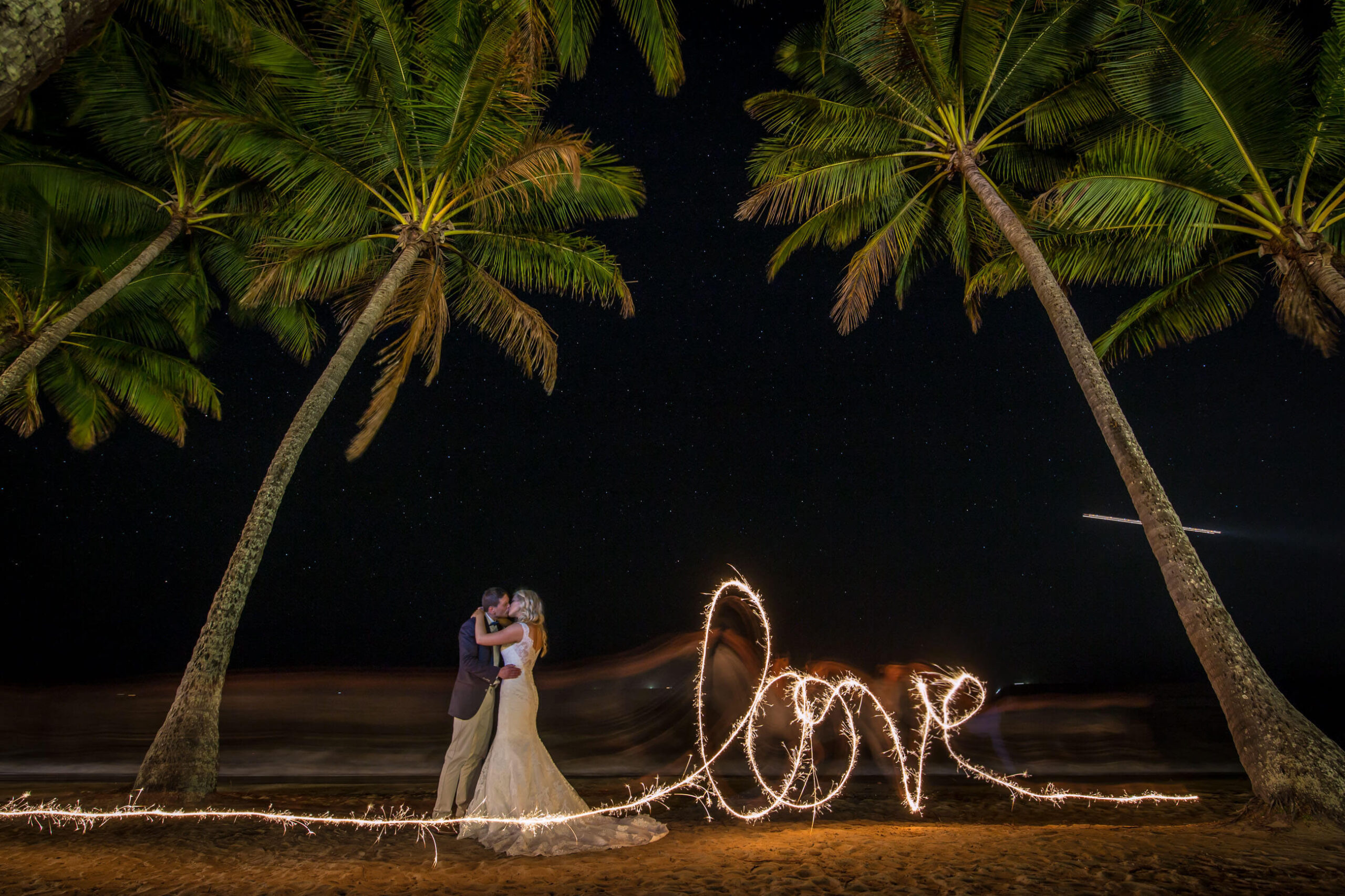 newlyweds kissing under coconut trees with “love” spelled with sparklers