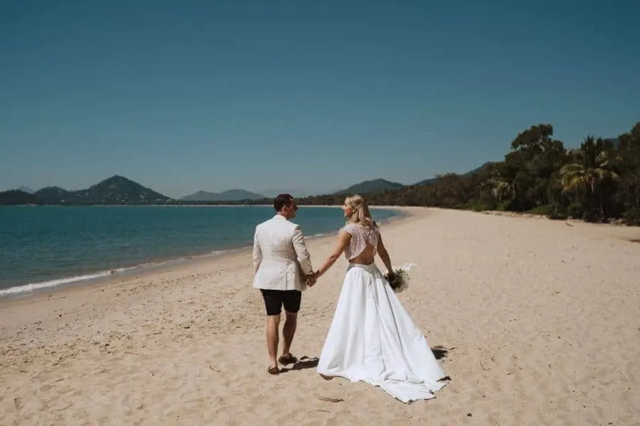 Groom wearing suit and shorts and bride wearing a gown taking a walk on the sandy beach