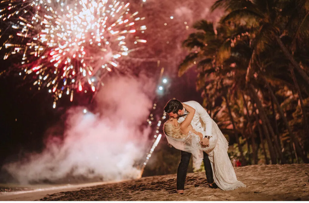 newlyweds kissing at the beach with fireworks in the background