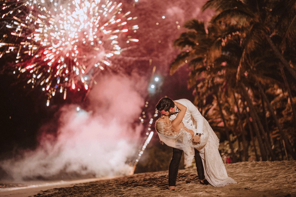 newlyweds kissing on the beach with fireworks as background